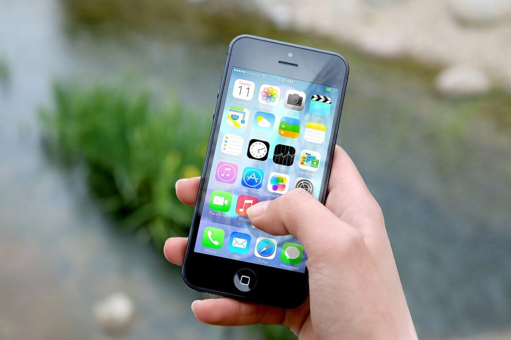 Hand holding a smartphone with various app icons on the screen, background blurred nature scene.