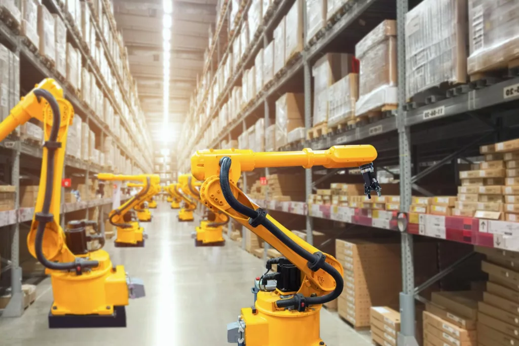 A series of yellow robotic arms in a well-lit warehouse aisle, with shelves stacked with boxed goods on either side.
