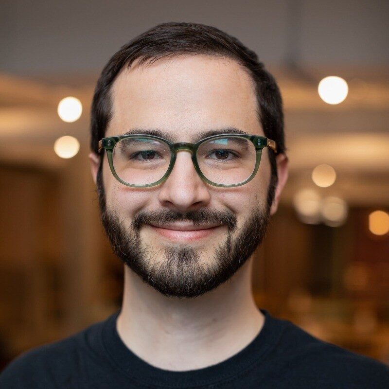 Headshot of Reid Robinson smiling warmly, wearing green-rimmed glasses and a dark shirt, set against a softly blurred indoor background with warm lighting.